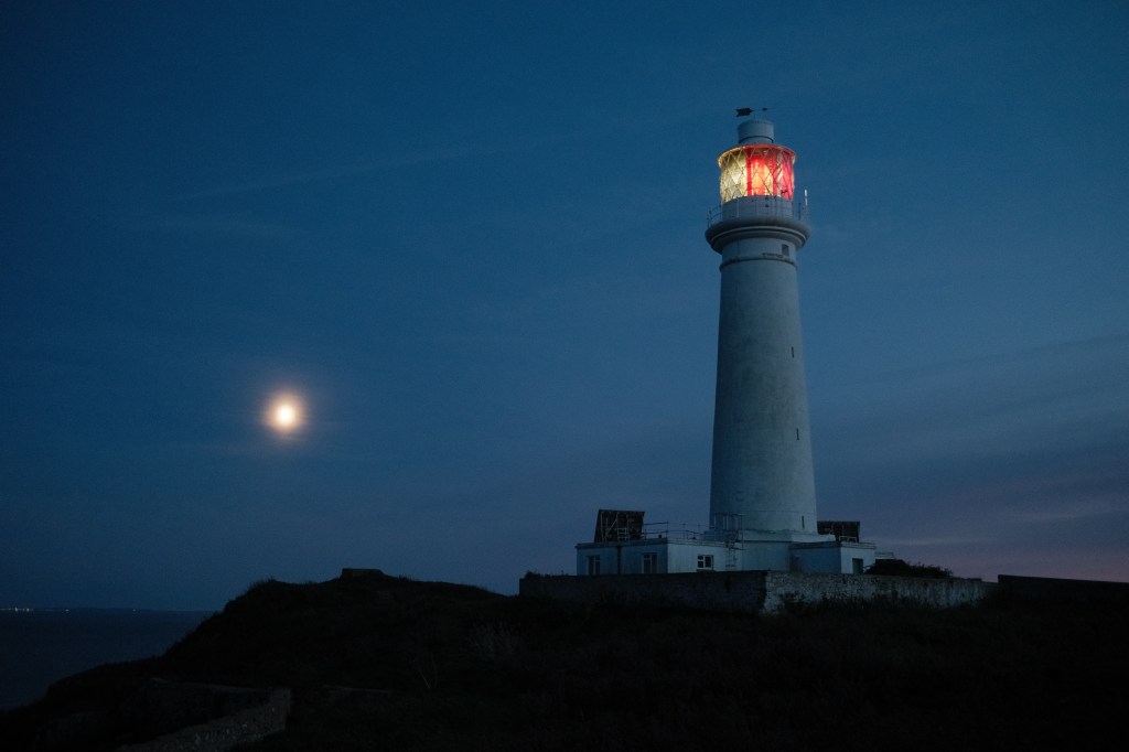 A lighthouse at night with the moon in the background. The light of the lighthouse is illuminated red and white.