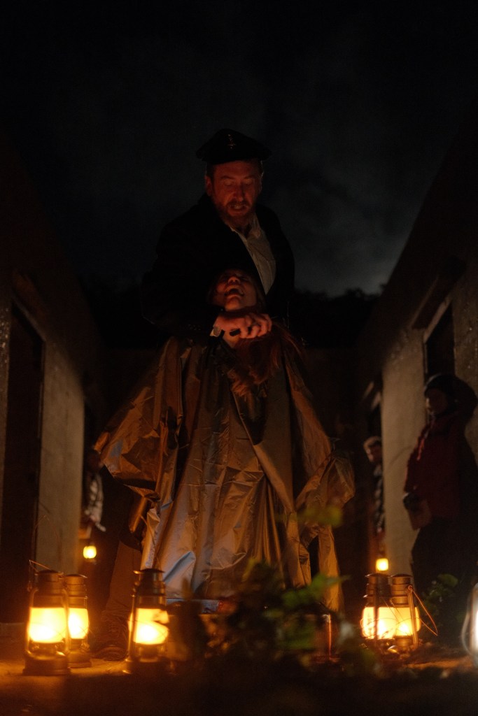 A man stood behind a woman with a knife to her neck. The woman is kneeling down beside a device, surrounded by lanterns.