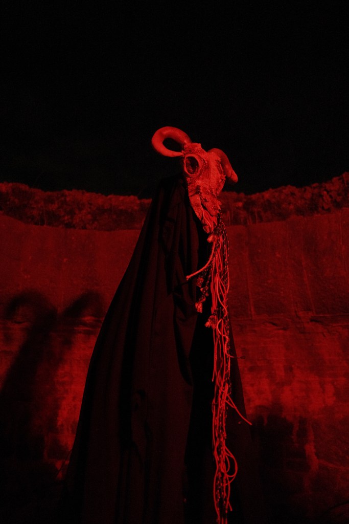 A tall monster at night. The monster has a head that looks like a sheep's skull. The picture is backlit in red.