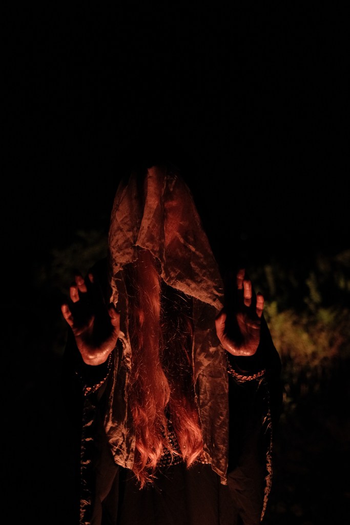 A witch holding out her hands. It is dark. Her face is covered.