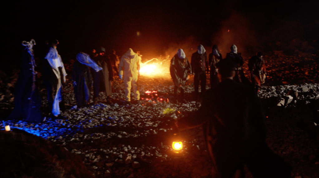 9 monsters stood on a rocky beach with a fire in the background. Sebastian is stood facing the ghosts.