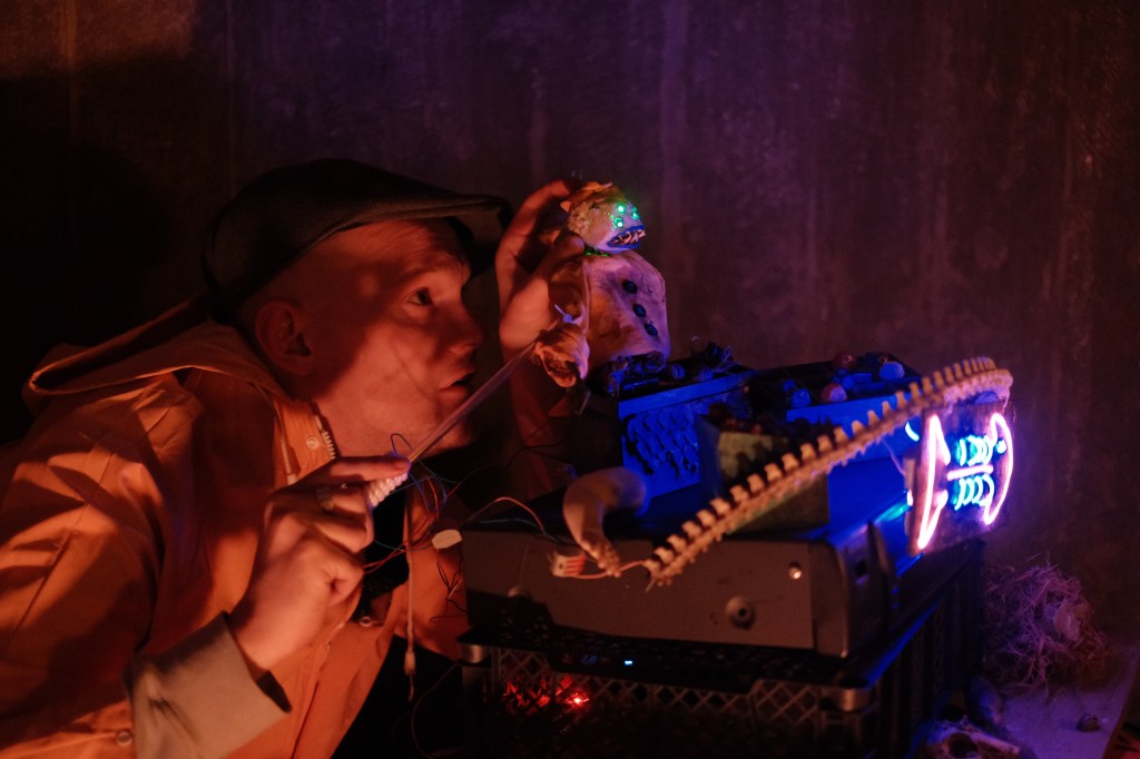 A man in a yellow fisherman's coat controls a small puppet that has three green eyes. There isa collection of wares.